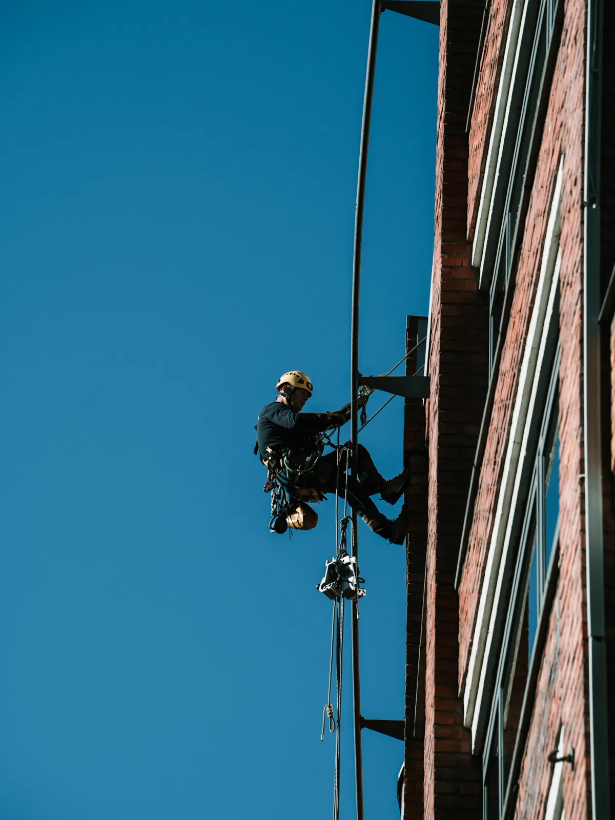 Rope access technician suspended beside a brick facade against a blue sky